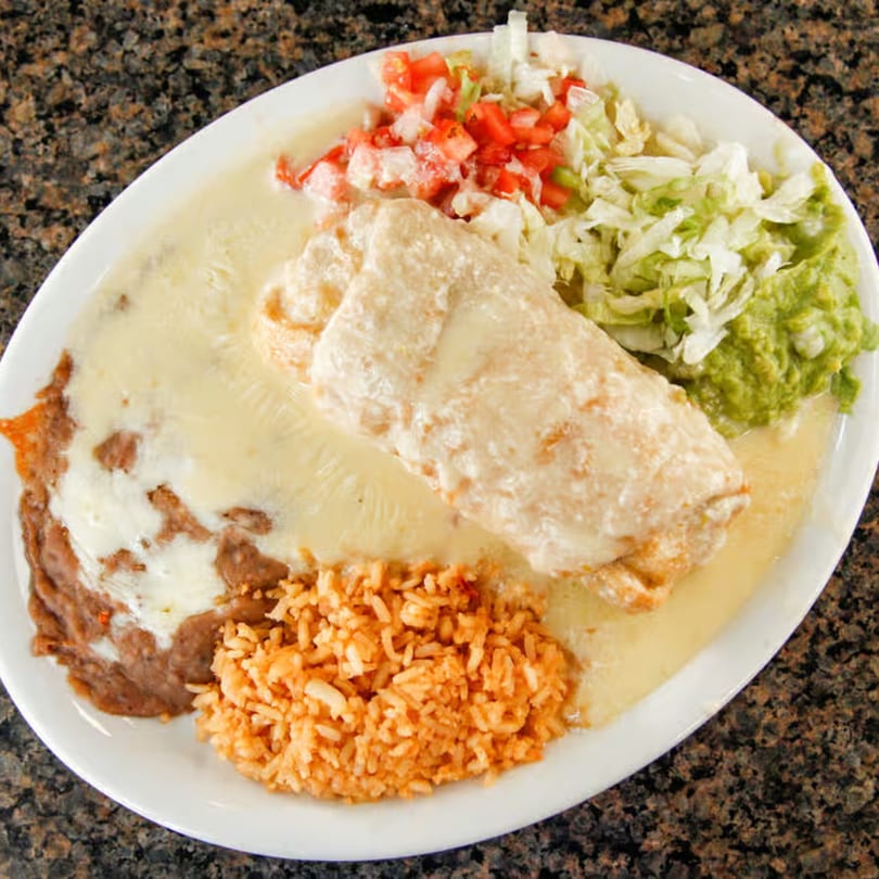 A cheese-covered burrito with refried beans, rice, lettuce, tomatoes, and guacamole on a white plate.