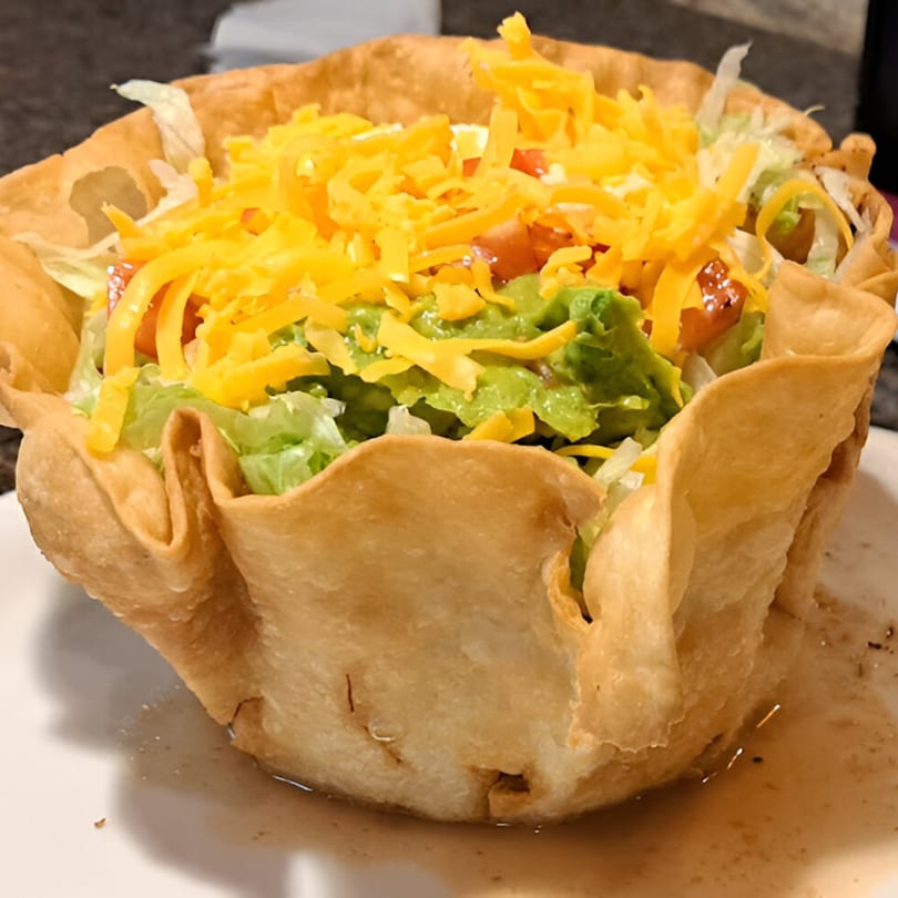 A taco salad in a crispy fried tortilla bowl, topped with lettuce, guacamole, tomatoes, and shredded cheese.